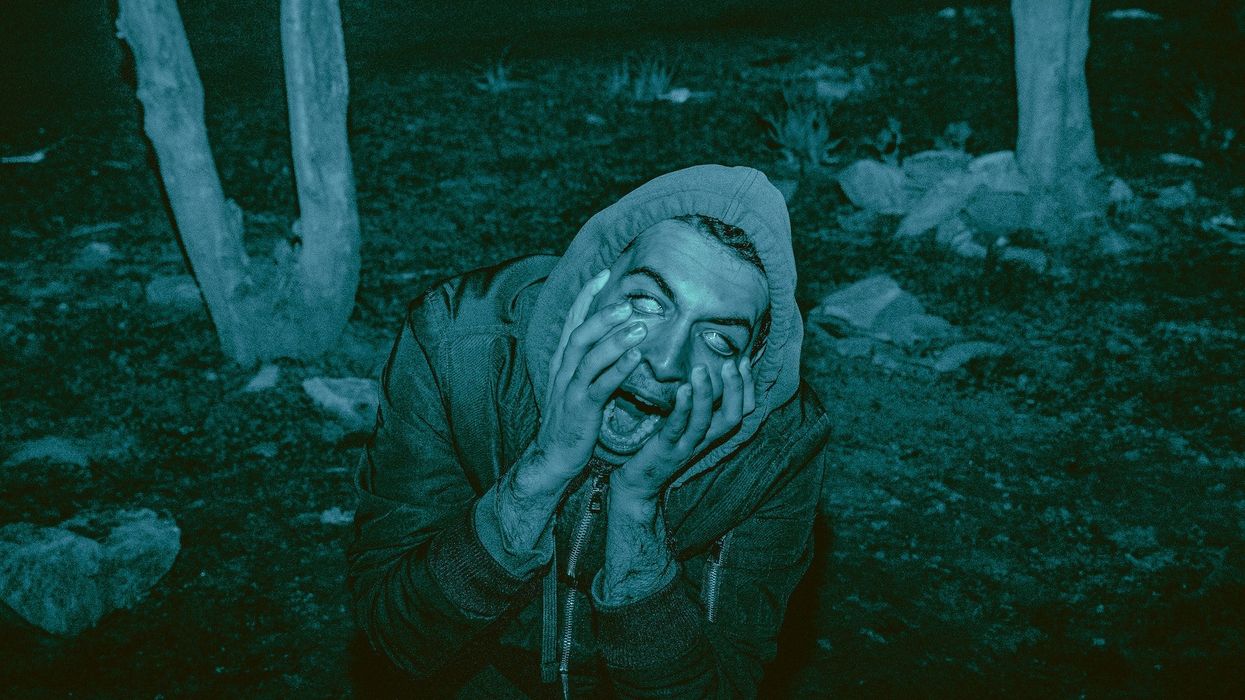 A young man in the woods is on his knees screaming, hands pulling at his face, with only the whites of his eyes showing.