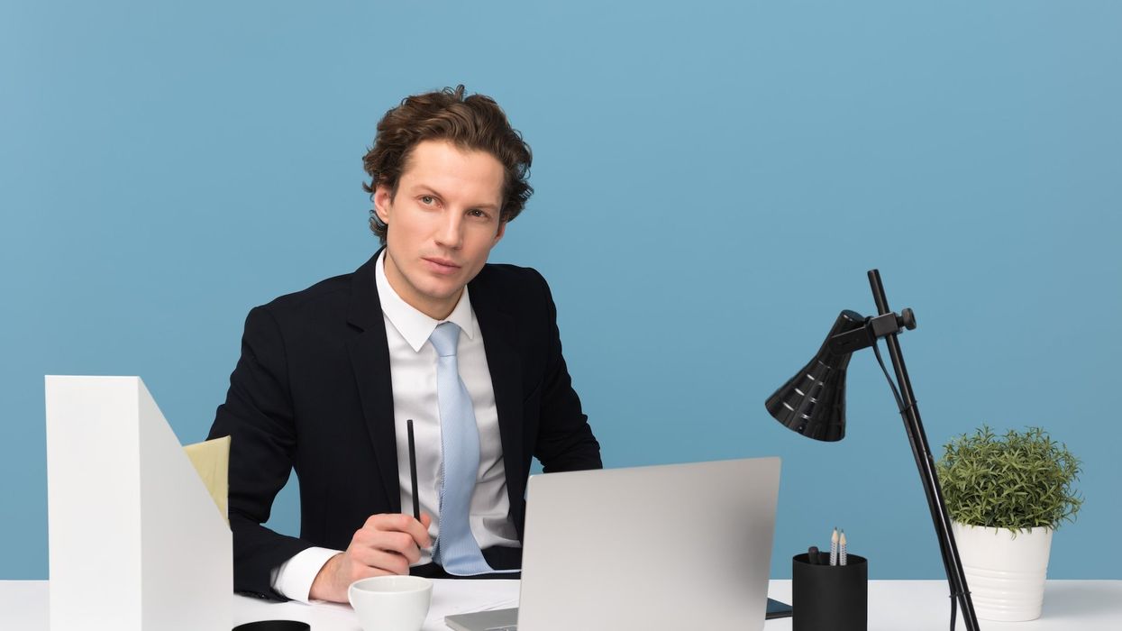 A young man sits at a desk in an office, thinking