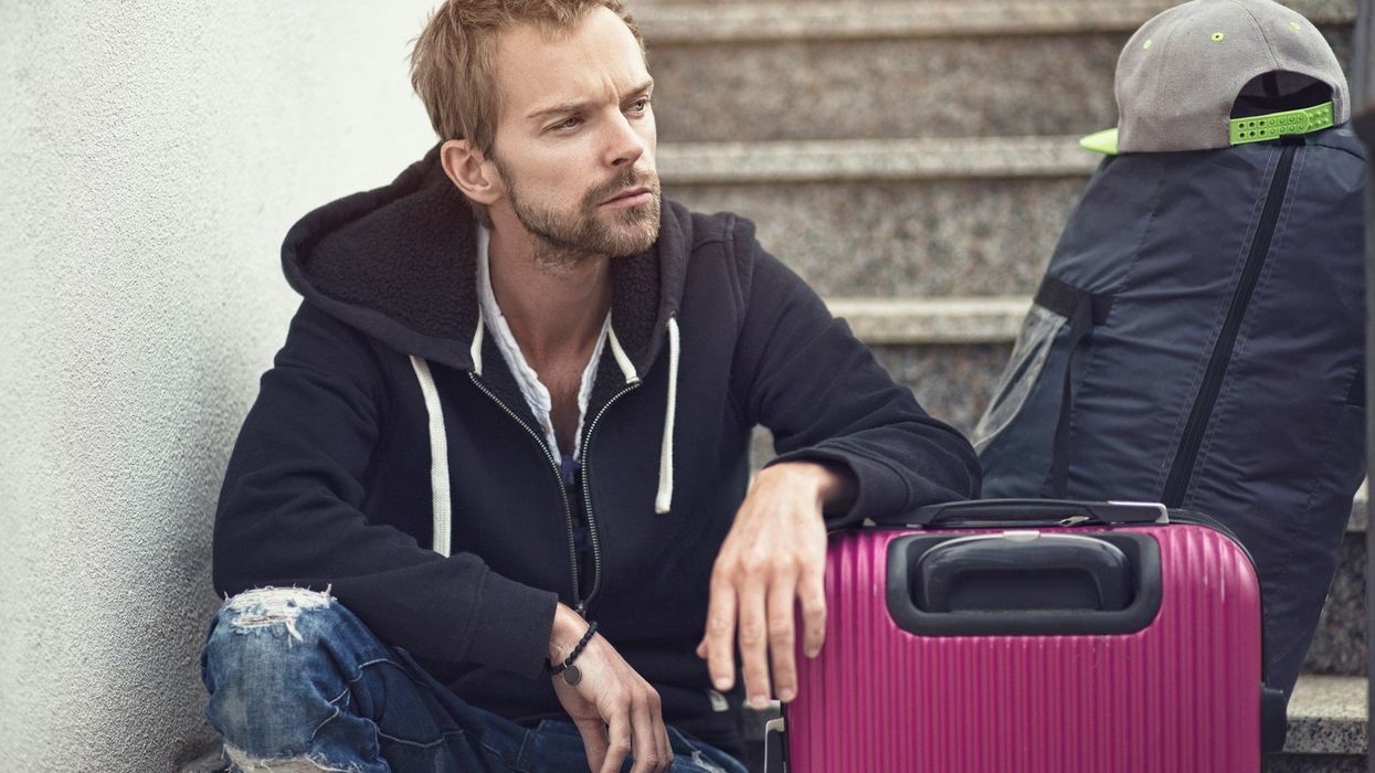 A young man sits on an outdoor staircase and looks longingly off into the distance. He has his luggage standing next to him.