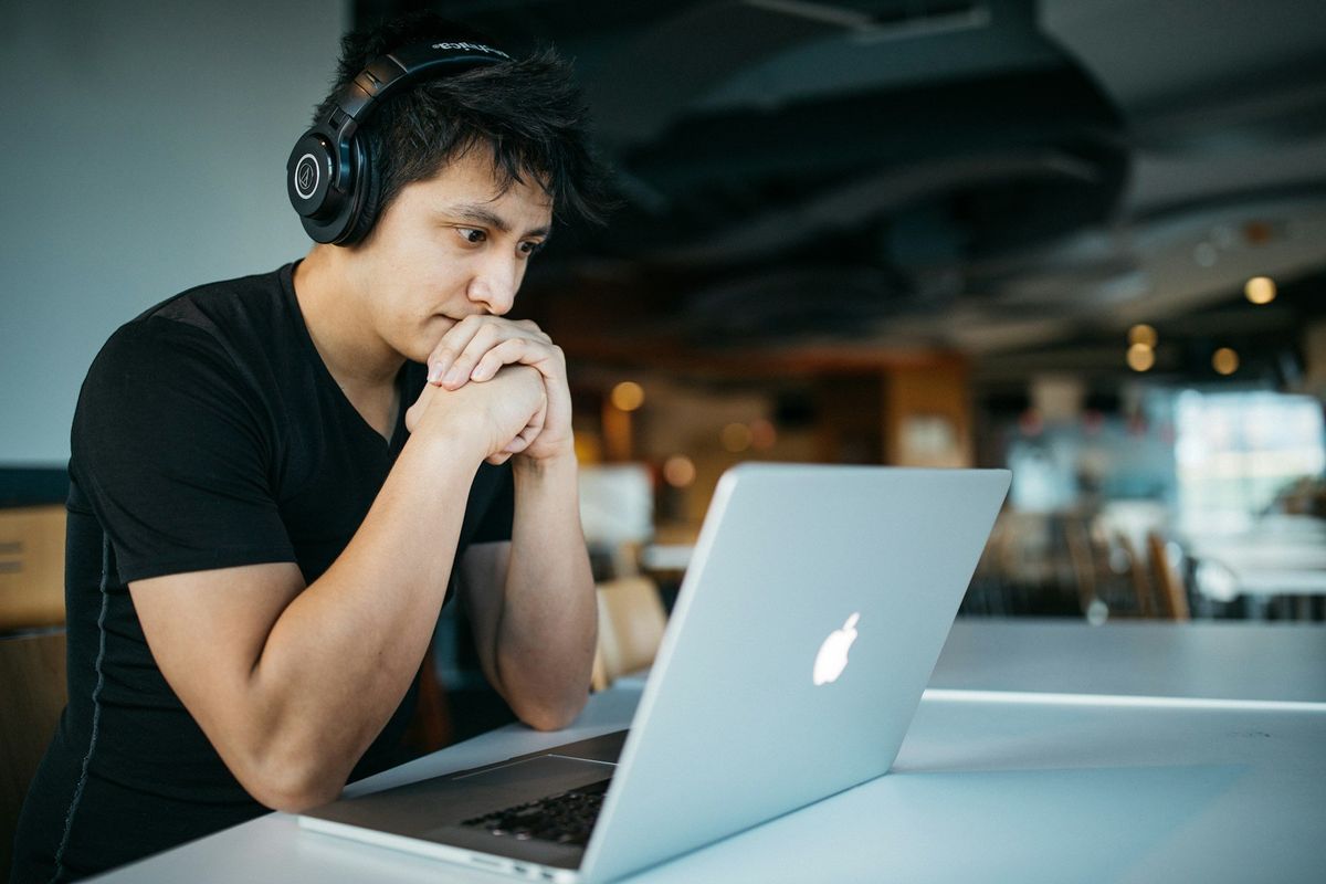 A young man, wearing headphones sits at a table looking into his computer. He seems slightly concerned.
