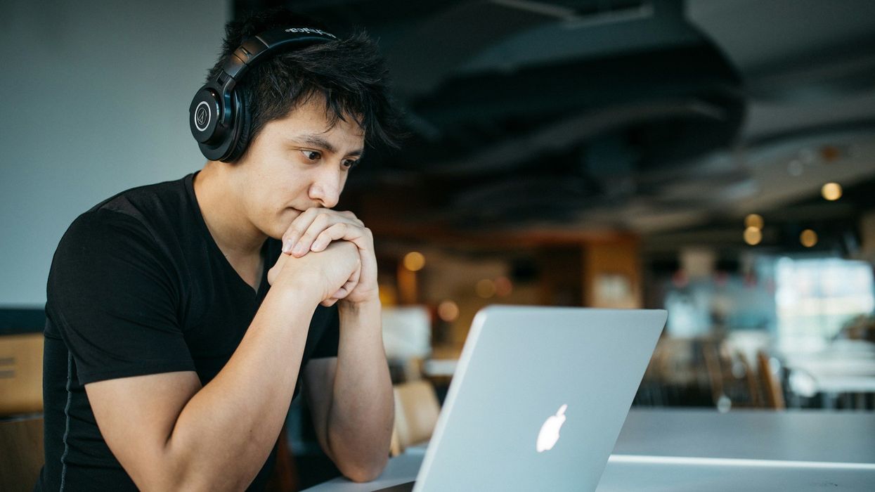 A young man, wearing headphones sits at a table looking into his computer. He seems slightly concerned.