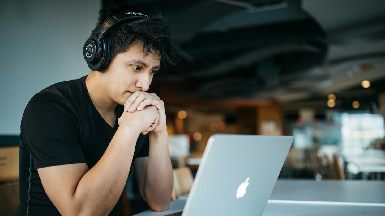 A young man, wearing headphones sits at a table looking into his computer. He seems slightly concerned.