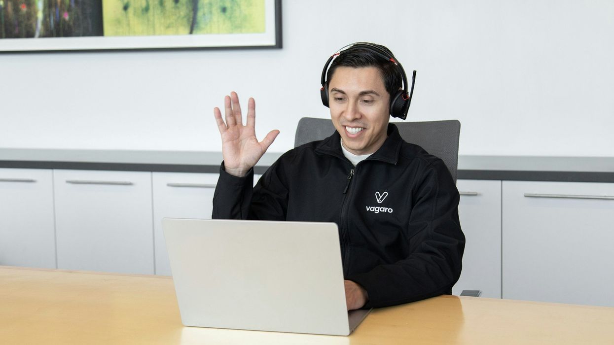 A young man with a huge smile sits at a desk, waving at someone on his laptop.