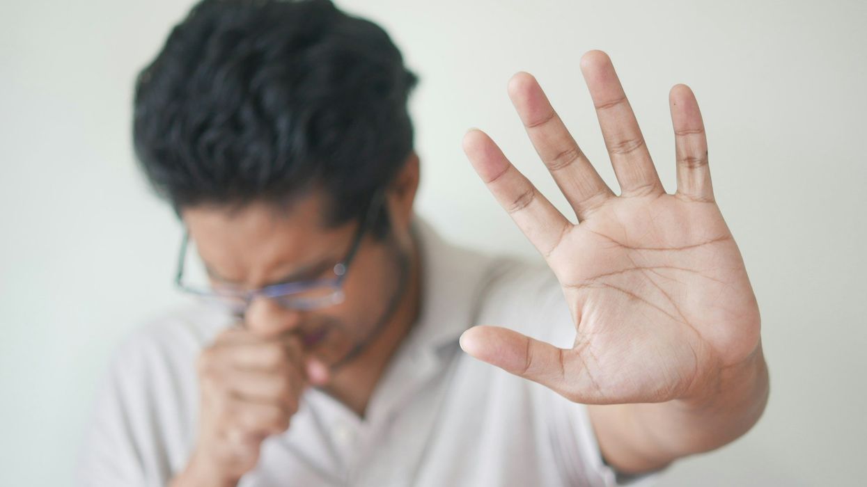 A young man with glasses is queasy. He holds his hand to his mouth and extends his other hand out to the camera signaling for someone to go away.