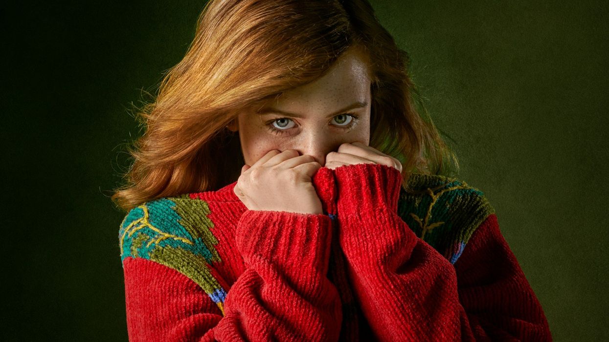 A young, pretty red-haired girl hides her mouth and looks sheepishly into the camera. She stands in front of a dark green background.
