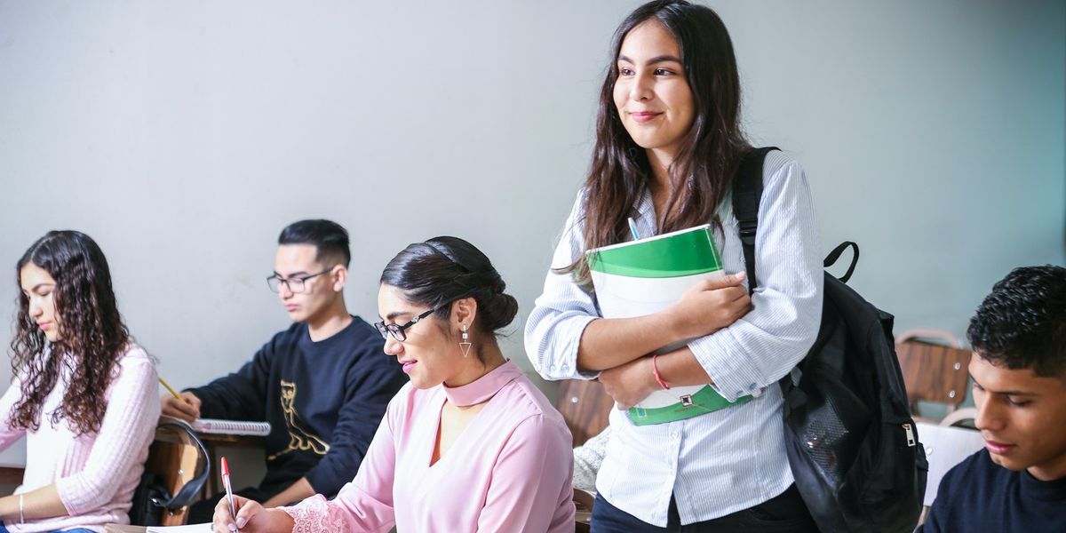 A young woman holding books, stands in a classroom smirking.