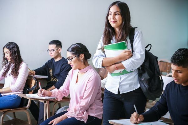 A young woman holding books, stands in a classroom smirking.