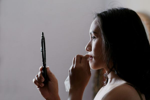 A young woman looks into a hand mirror while she wipes her mouth with a napkin.