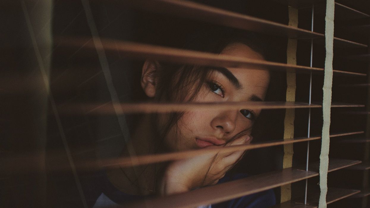 A young woman looks longingly out a window, through blinds