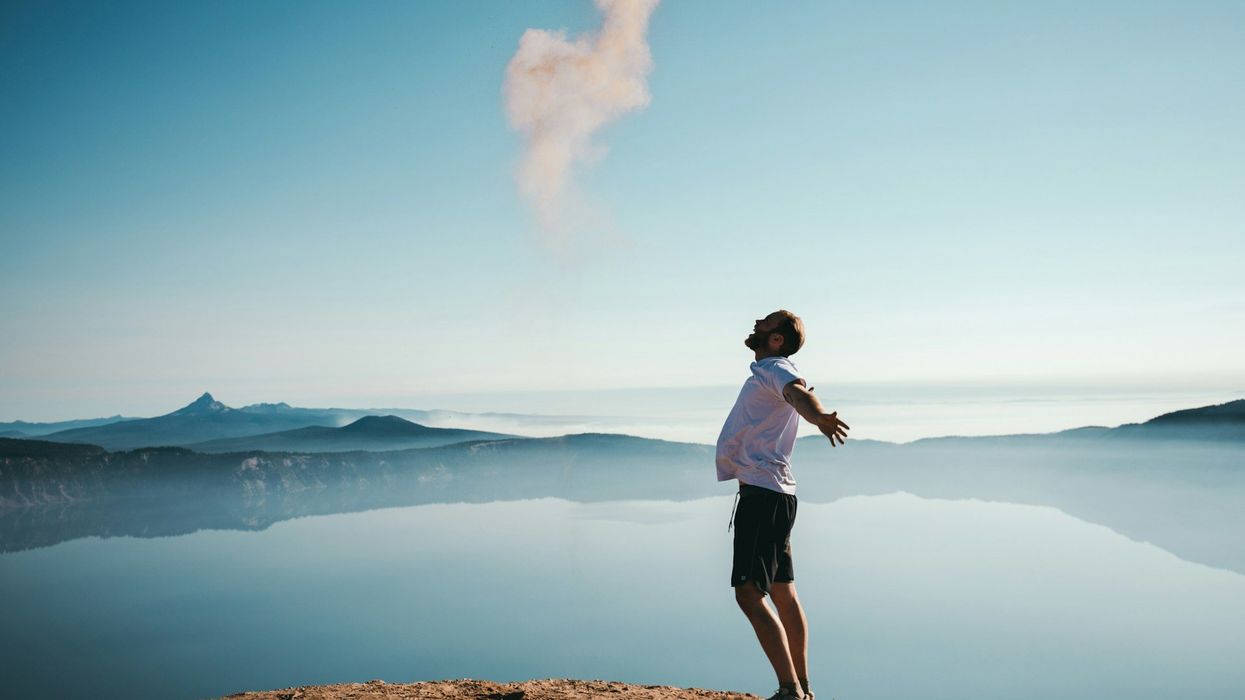 A younger man stand on top of a mountain with his arms outreached and his face looking to the sky. It's a beautiful day and lakes and mountains are the backdrop.