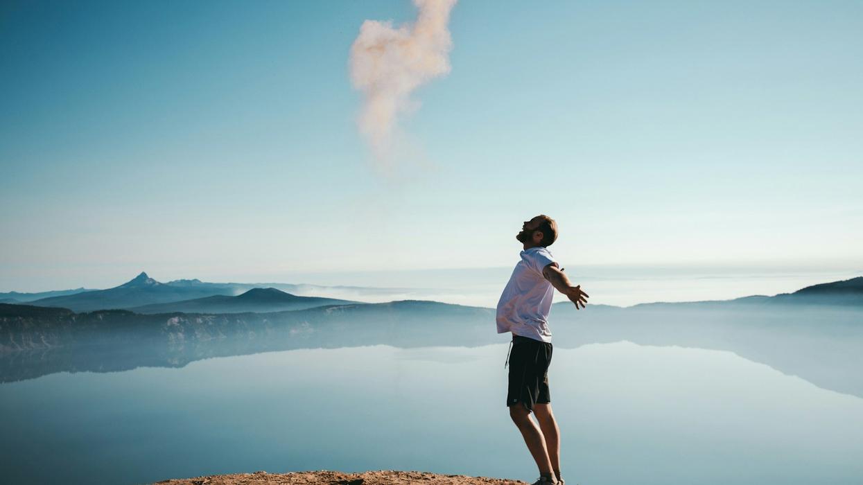 A younger man stand on top of a mountain with his arms outreached and his face looking to the sky. It's a beautiful day and lakes and mountains are the backdrop.