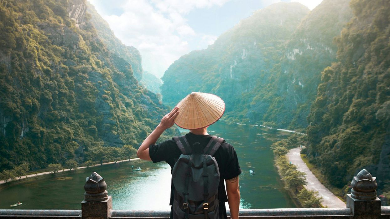 Back shot of a man wearing a straw Kasa, as he looks out at majestic mountains and a river.