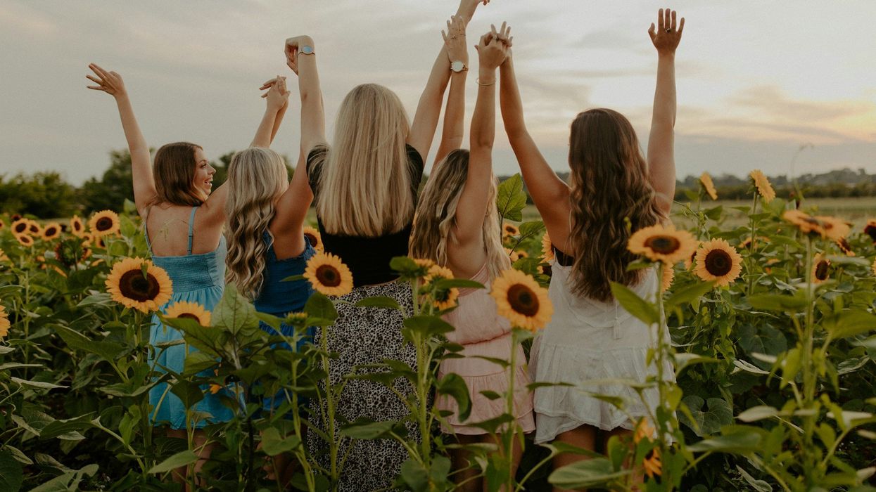 Back shot of five young, carefree female friends stand in a field of tall sunflowers clasp hands and raise their arms to the sky.