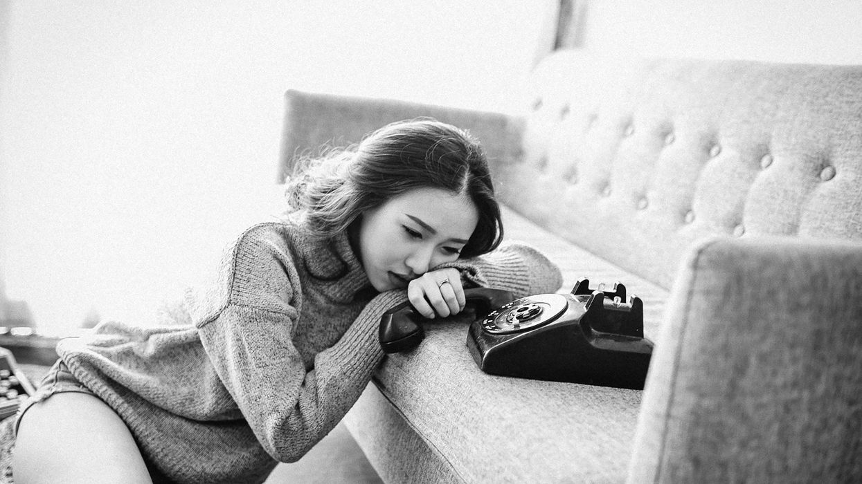 Black and white photo. A sullen young woman sits on the floor as she lies her head on her arm while holding a phone receiver.