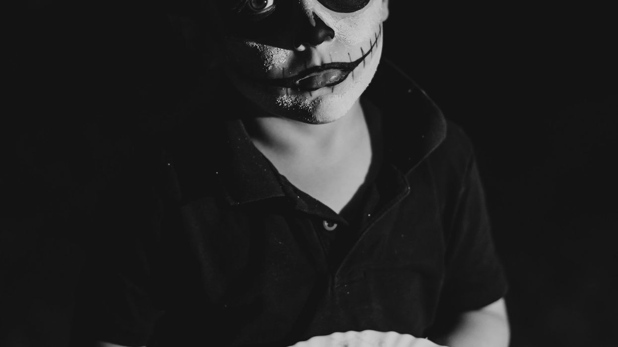 Black and white photo of a young boy. His face is covered in white paint, his eyes have painted in black circles and fake stitches are painted on.