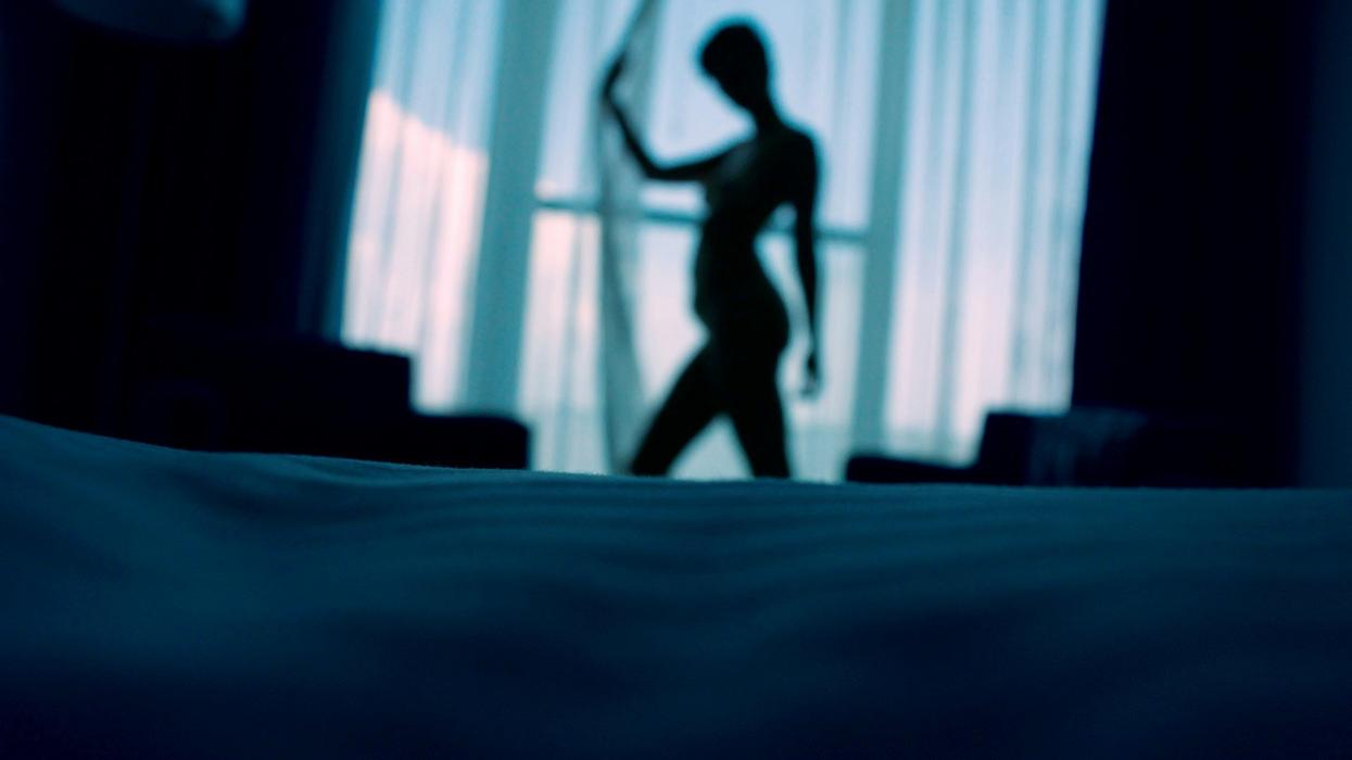 Blurry silhouette shot of a woman standing against a window, pulling a curtain back. The picture has a blue tint.
