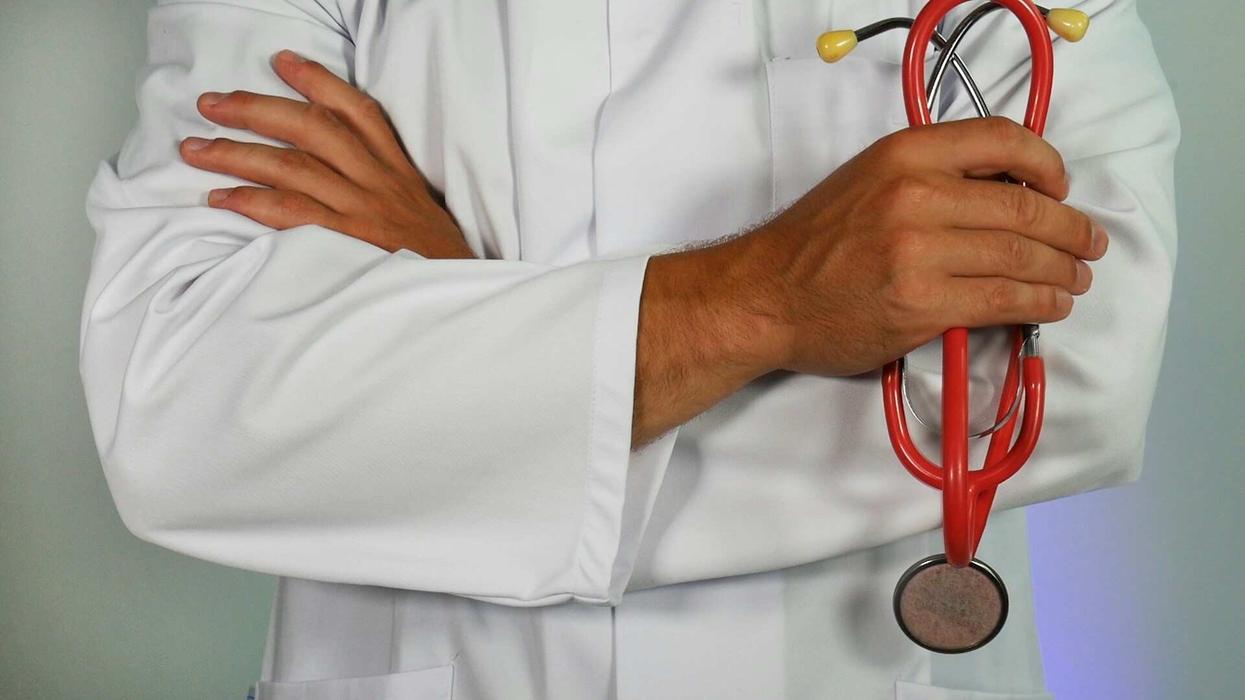Close-up, mid-section shot of an unidentifiable male doctor's body. In his right hand he holds a red stethoscope.