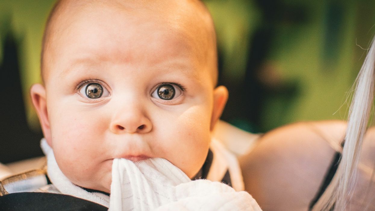 Close-up of a baby's face. Their eyes are wide in surprise. They suck on a piece of cloth.