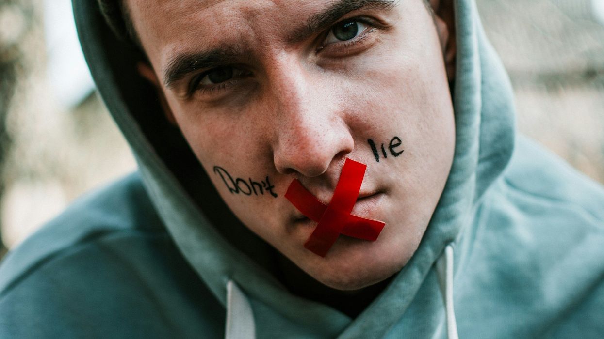 Close-up of a serious looking young man's face, "Don't lie" is written on his cheeks and an X made of red tape is over his mouth. He is wearing a light blue hoodie.