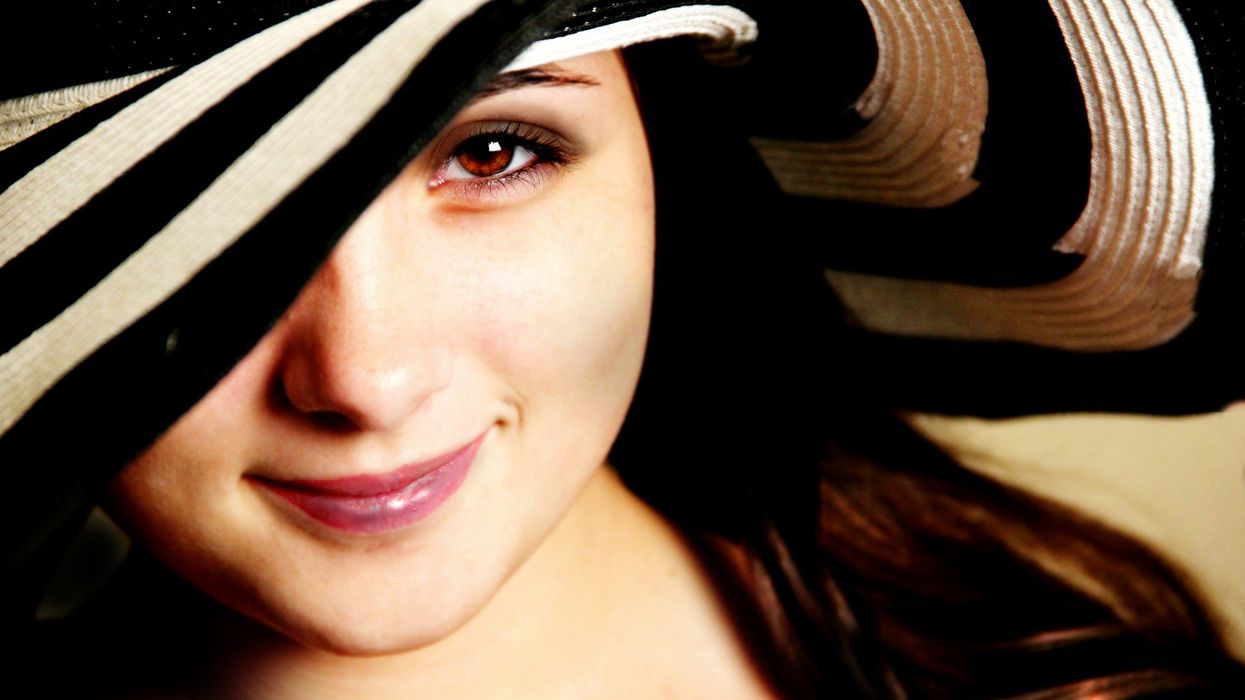 Close-up shot of a beautiful young woman looking coyly into the camera. She wears a large black and white beach hat.