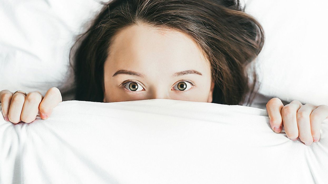 Close-up shot of a brunette woman's worried eyes peering out of white sheets, looking directly into the camera.