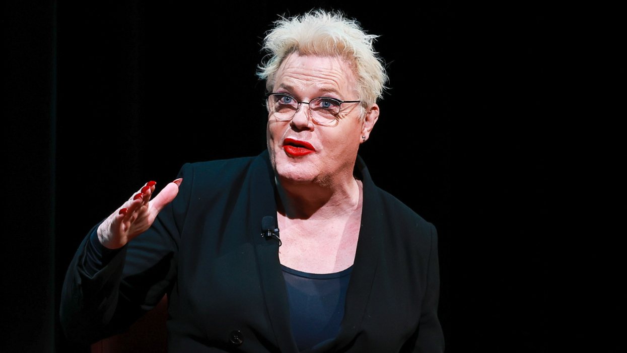 Comedian Suzy Eddie Izzard sits on a stage in front of a black background. She is in the middle of speaking and is gesturing with her right hand.