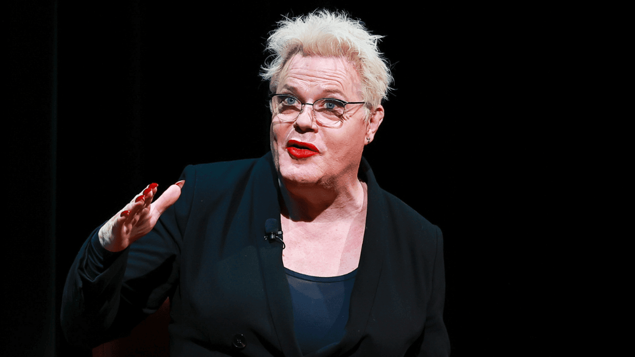 Comedian Suzy Eddie Izzard sits on a stage in front of a black background. She is in the middle of speaking and is gesturing with her right hand.