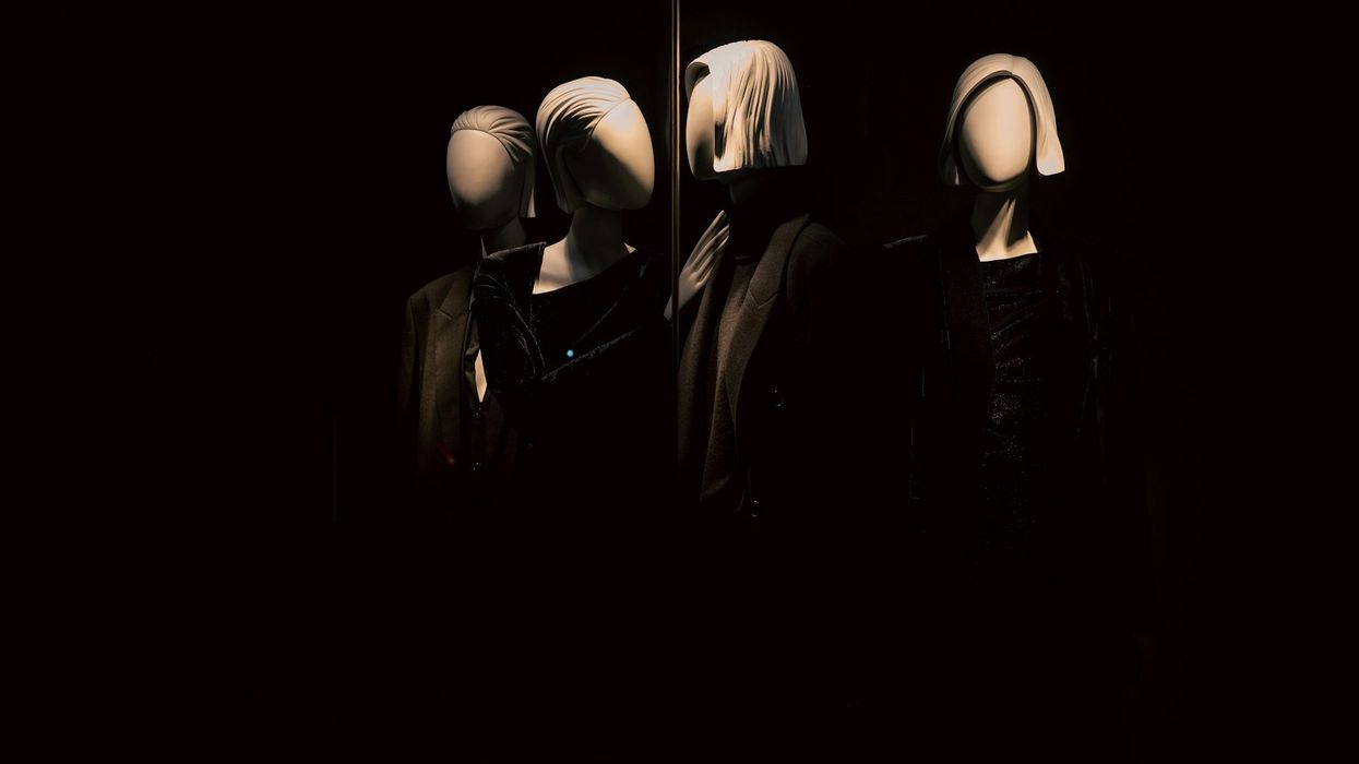 Dark shot of faceless mannequins positioned toward a mirror in an all black room. Each wears the same drab, black robe and white headpiece.