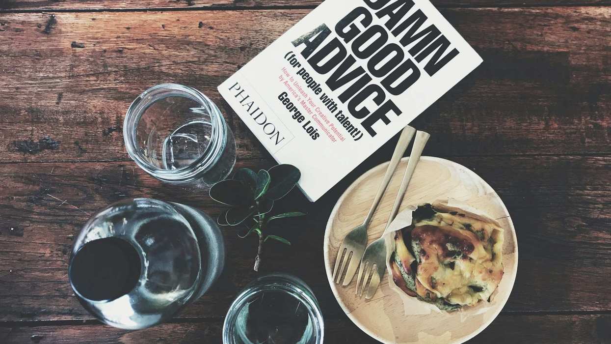 Downward shot of a book titled "DAMN GOOD ADVICE" with a plate of food and glass for water next to it. It all sits on a wooden table.