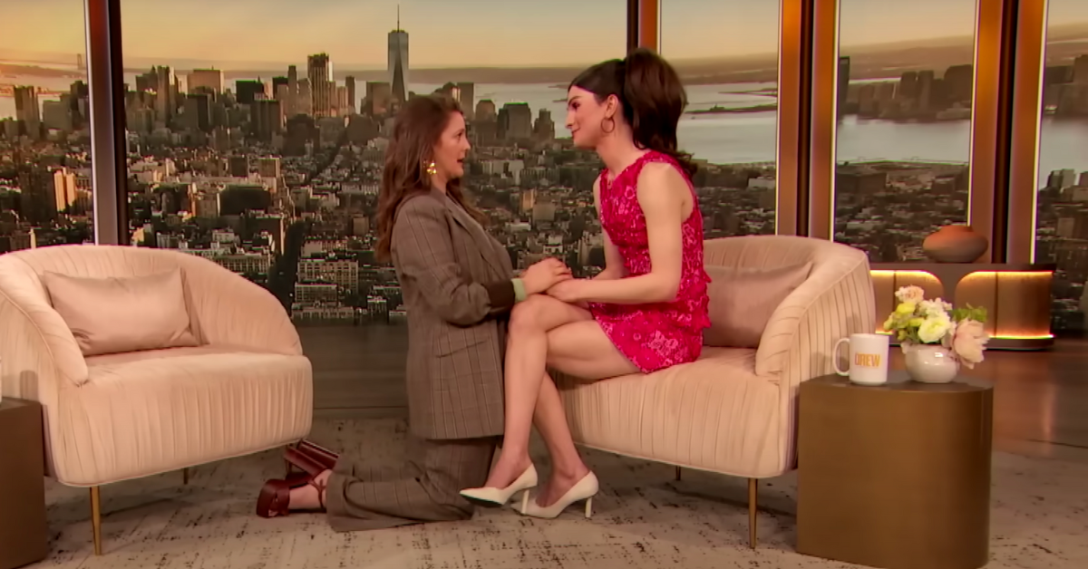 Drew Barrymore kneels in front of Dylan Mulvaney, holding her hands as the two have an emotional conversation