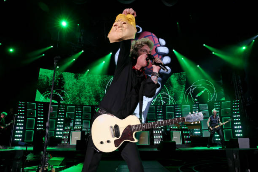 Green Day performing with Billie Joe Armstrong holding a Trump mask with "idiot" written on the forehead