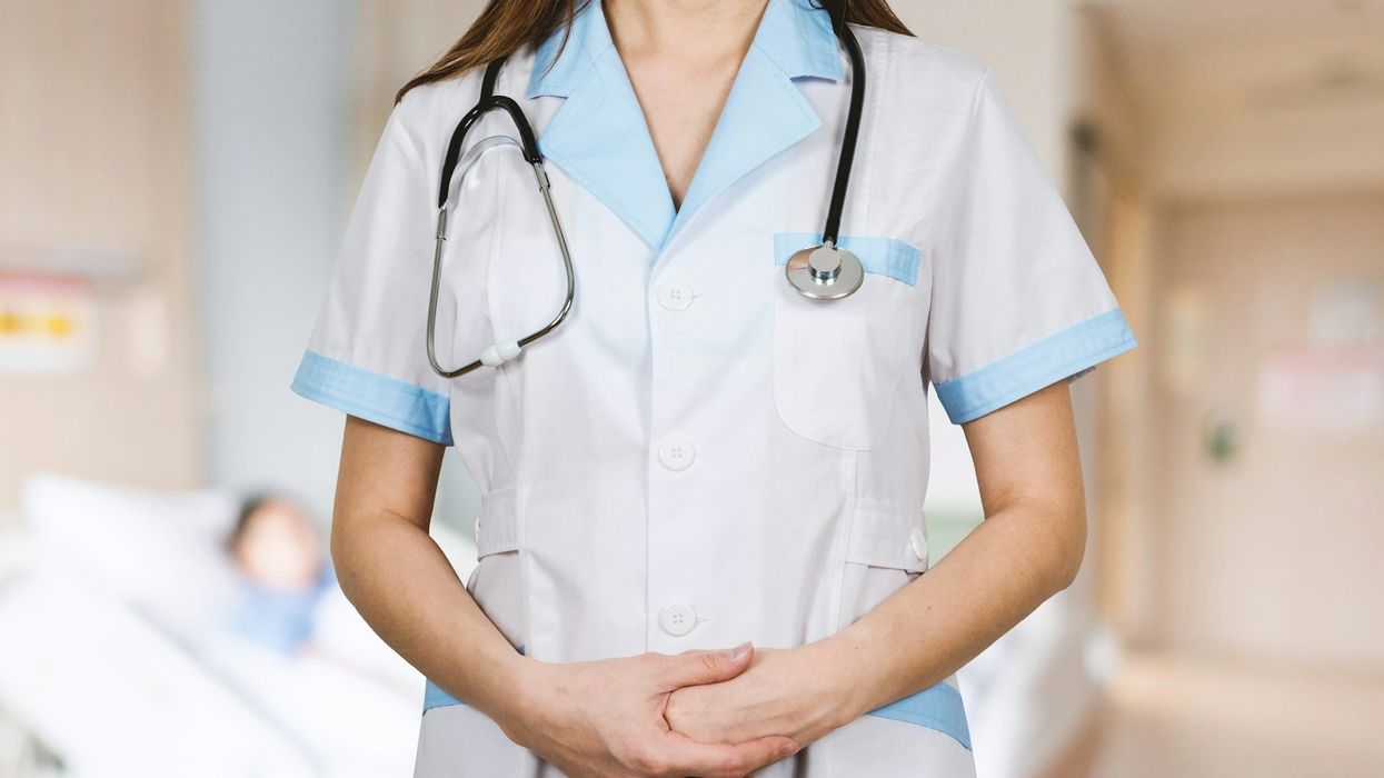 Headless, mid-shot of long, brown-haired nurse looking forward. She has a stethoscope around her neck and blurred out patient's face is in the background.