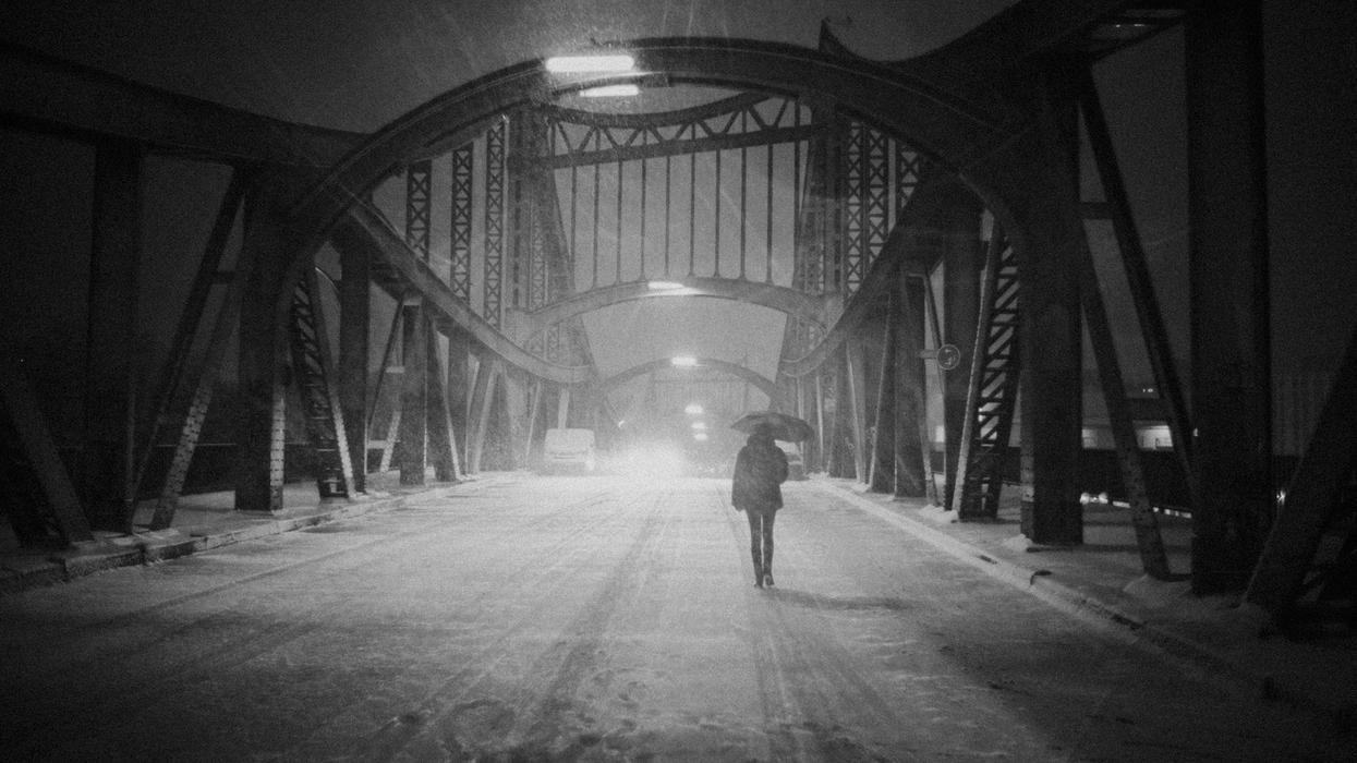 In a black and white photo a woman walks alone on a desolate bridge, in the snow, holding an umbrella