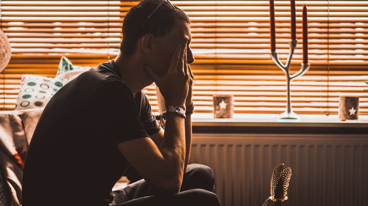 In a sepia toned photo, a young man sits on a couch, sad and rubbing his eyes.