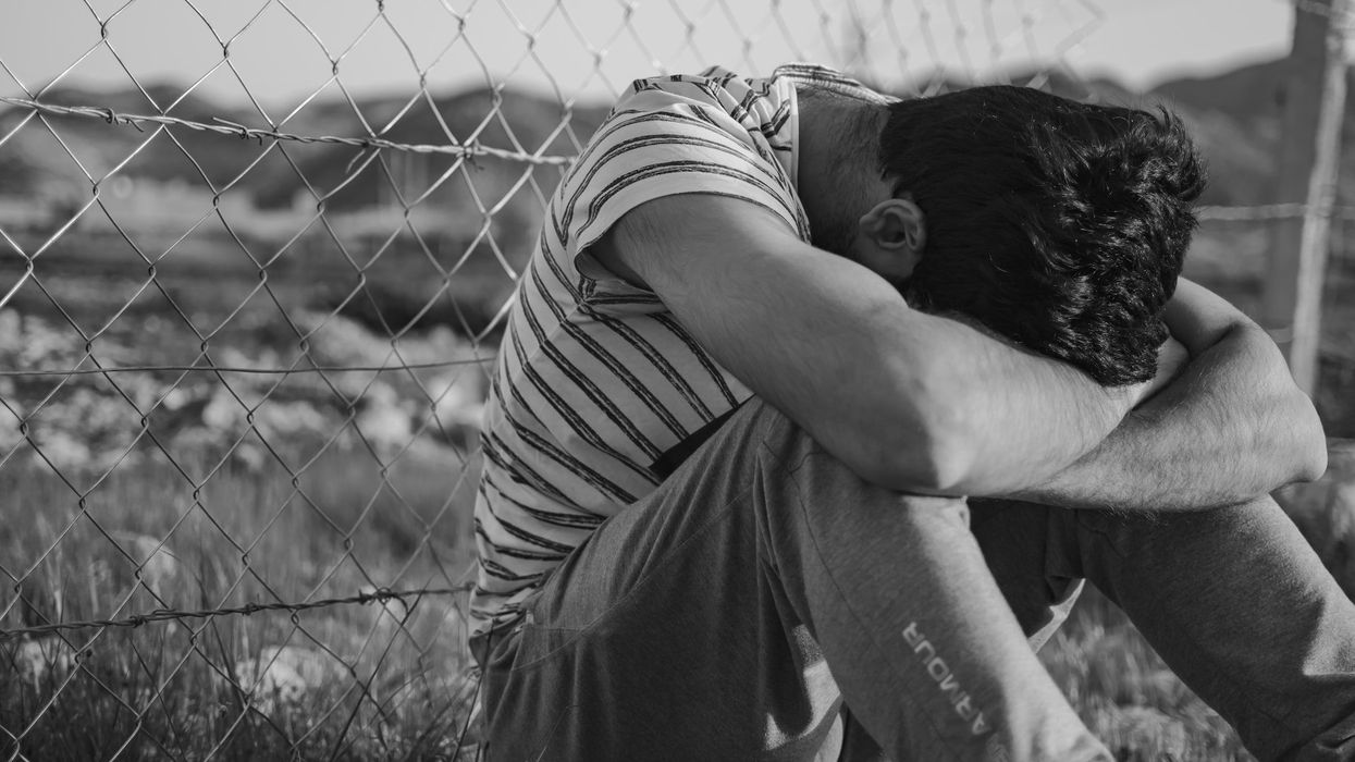 Man in a black and white stripe t-shirt sits along a chain link fence, with his head in his knees