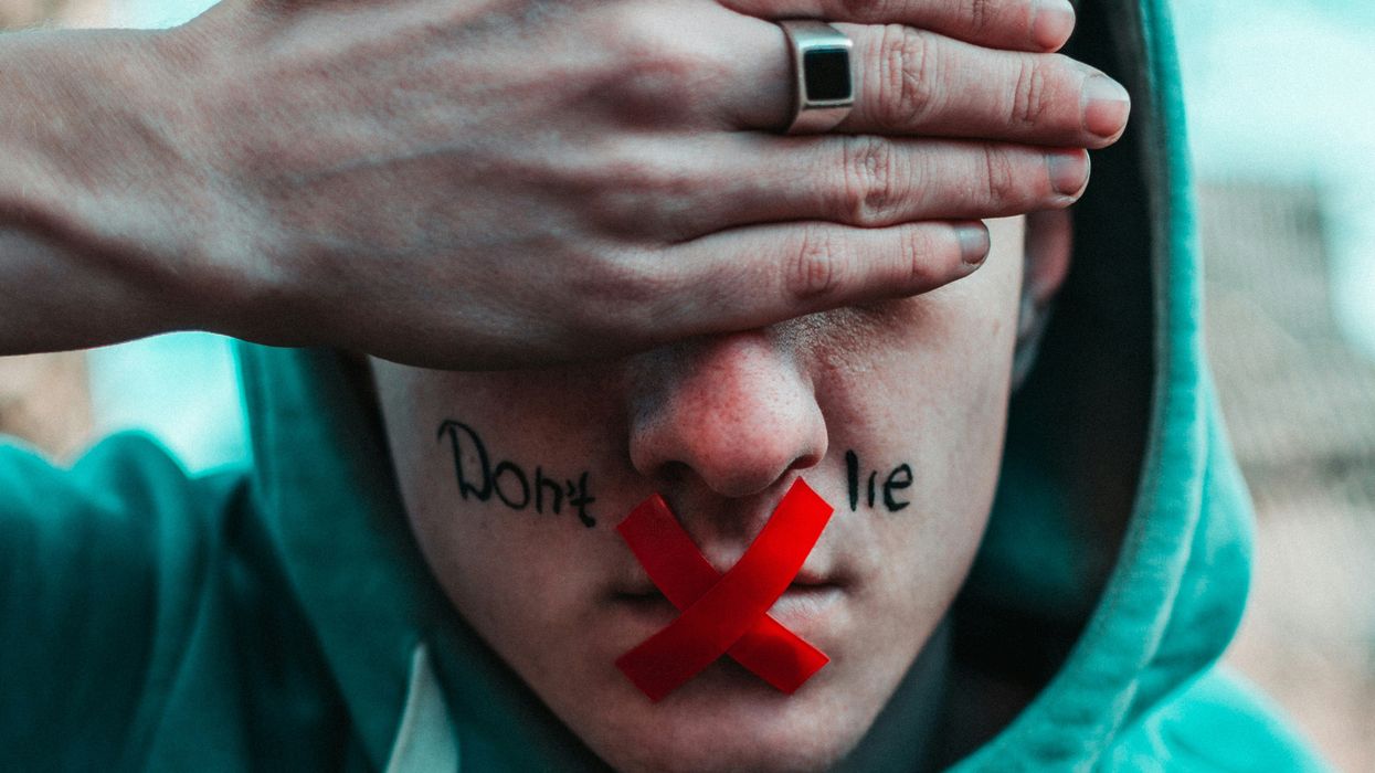 man in teal hoodie with don't lie written on face, red tape on mouth and hand over eyes
