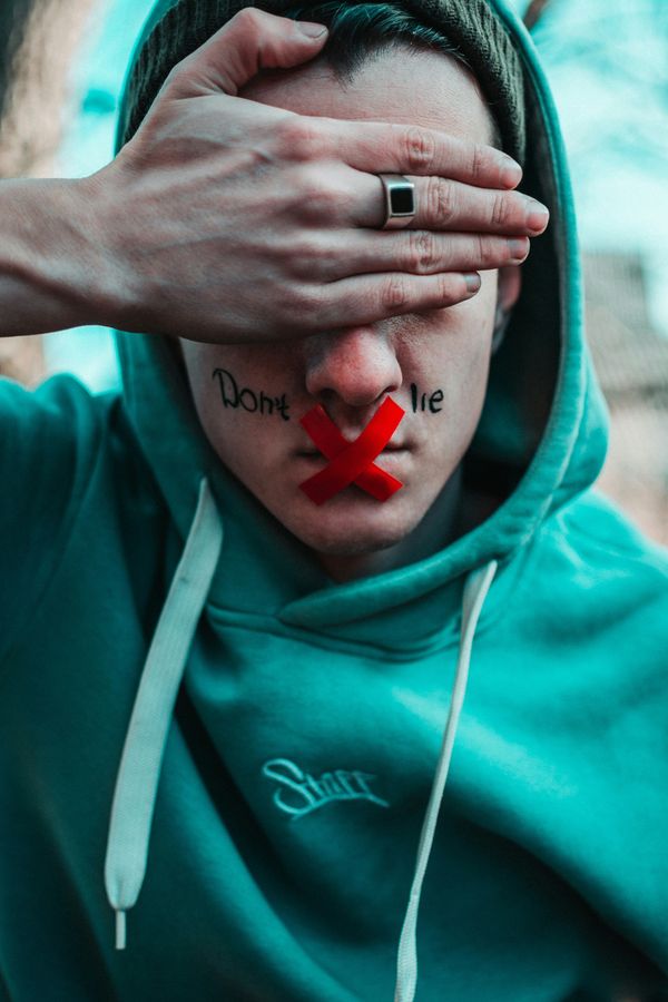 man in teal hoodie with don't lie written on face, red tape on mouth and hand over eyes