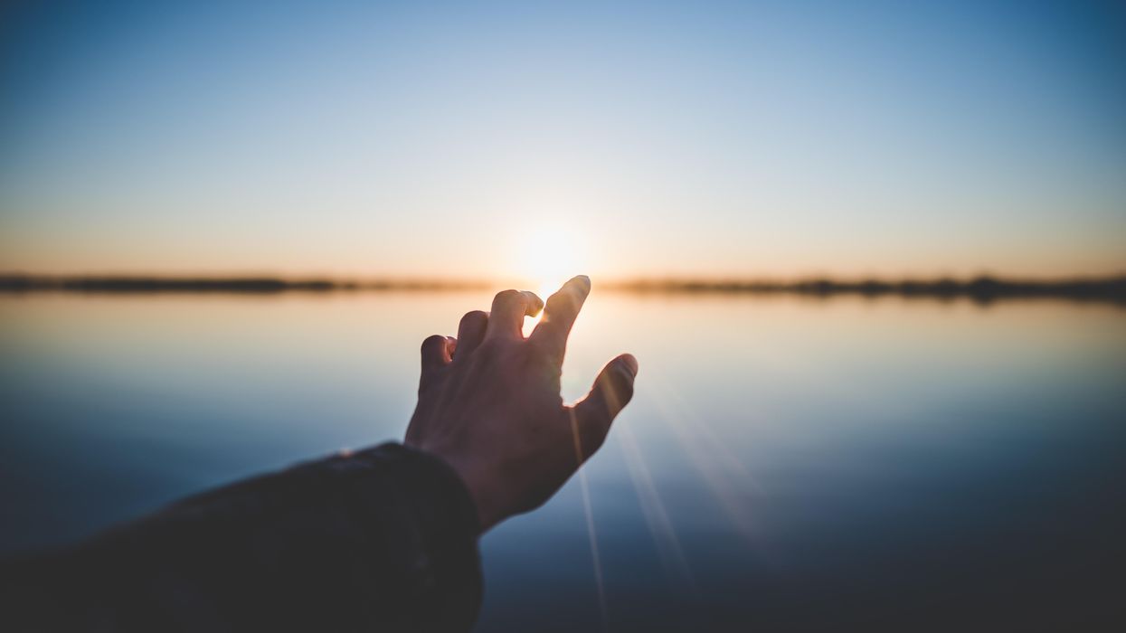 man's hand reaching toward the setting sun over a lake