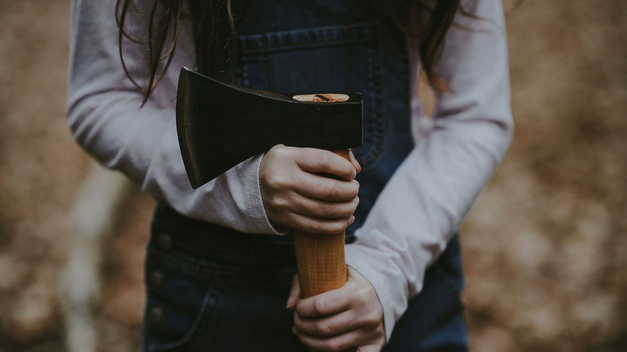 Mid shot a young girl's torso. She wears denim overalls and holds an axe.