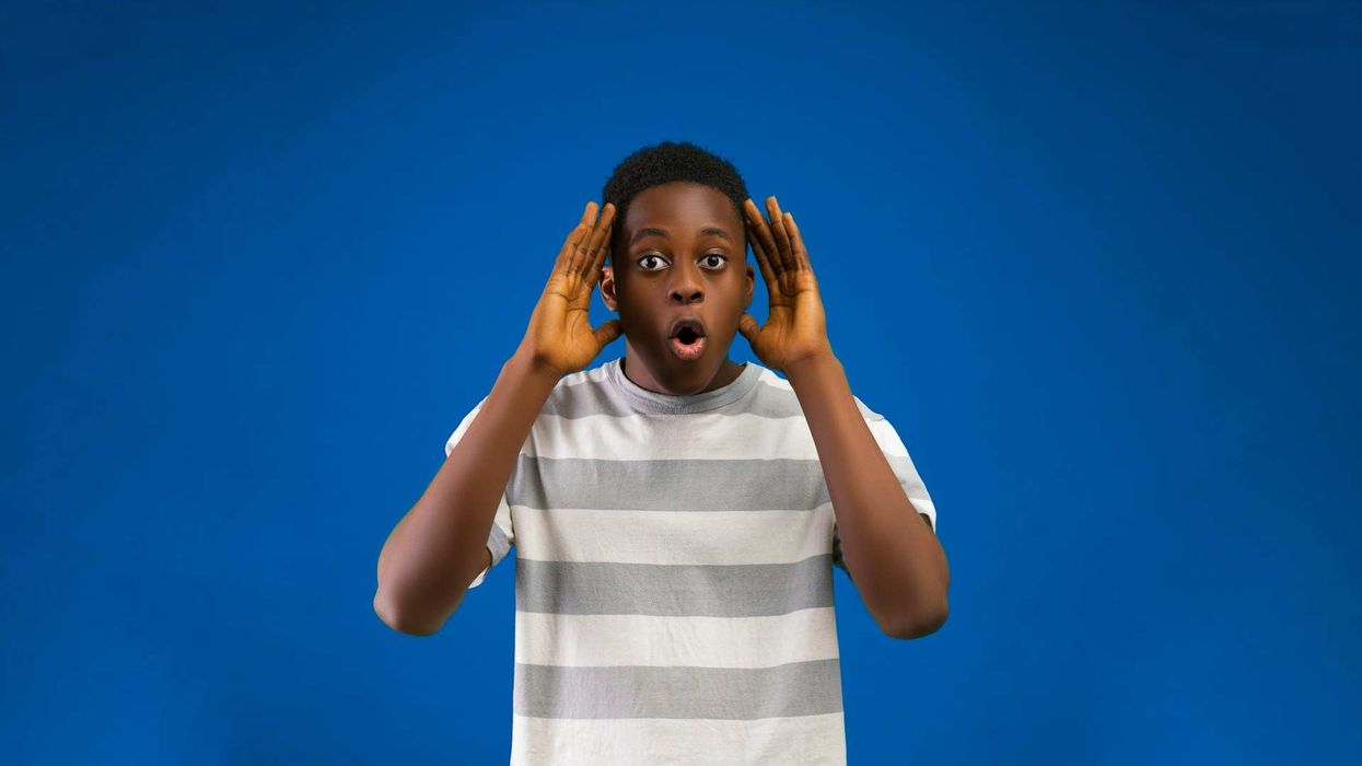Mid-shot of a teenage boy in a gray and white t-shirt, standing against a blue wall. His hands are open on both sides of his face. He is in shock.