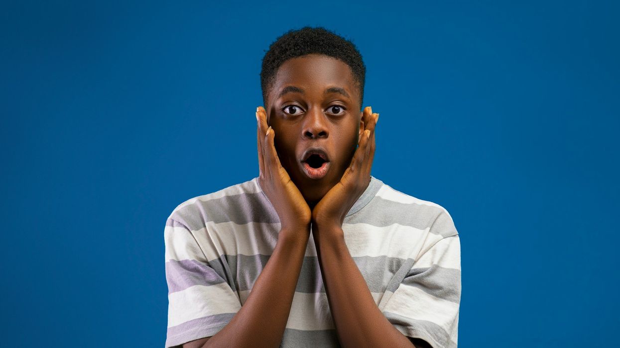 Mid-shot of a young man holding his face in shock. He stands in front of a blue wall.