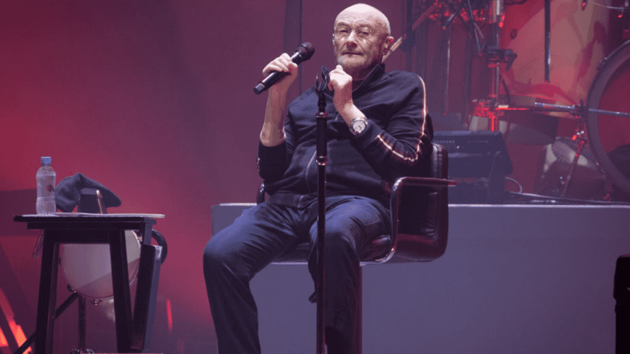 Phil Collins sitting on a chair on stage holding a microphone