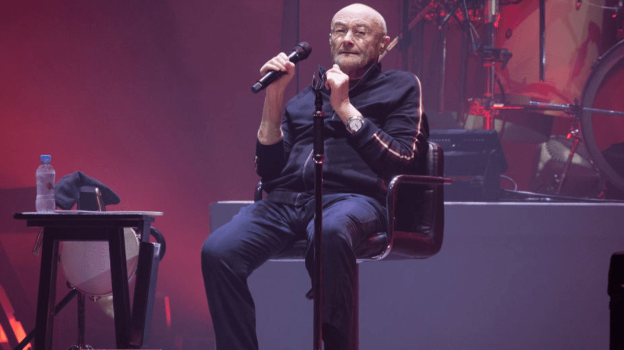 Phil Collins sitting on a chair on stage holding a microphone