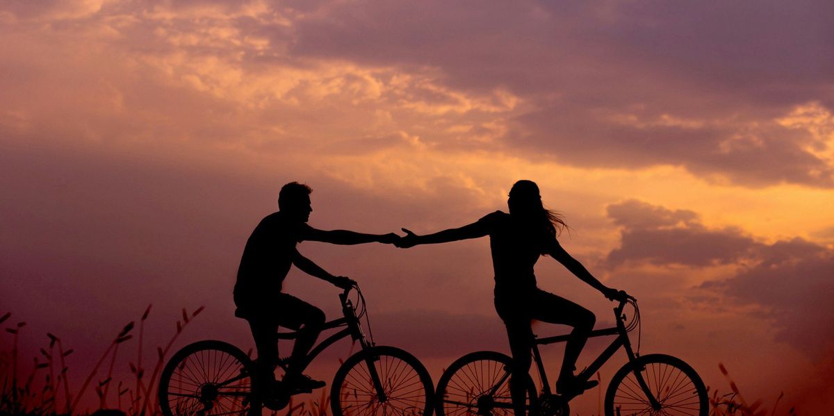 Photo of a couple in shadow, reaching out for one another as they ride bikes against the backdrop of sunset.