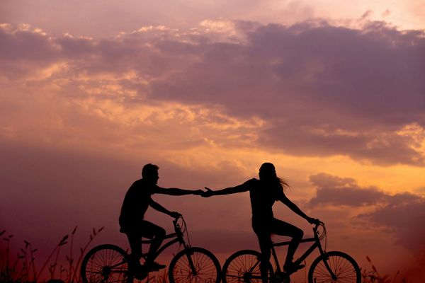 Photo of a couple in shadow, reaching out for one another as they ride bikes against the backdrop of sunset.
