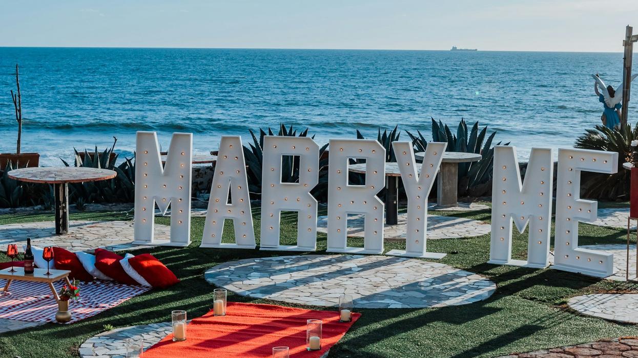 Photo of a massive sign saying "Marry Me." It's white with light bulbs. It sits on a fancy lawn overlooking an ocean.