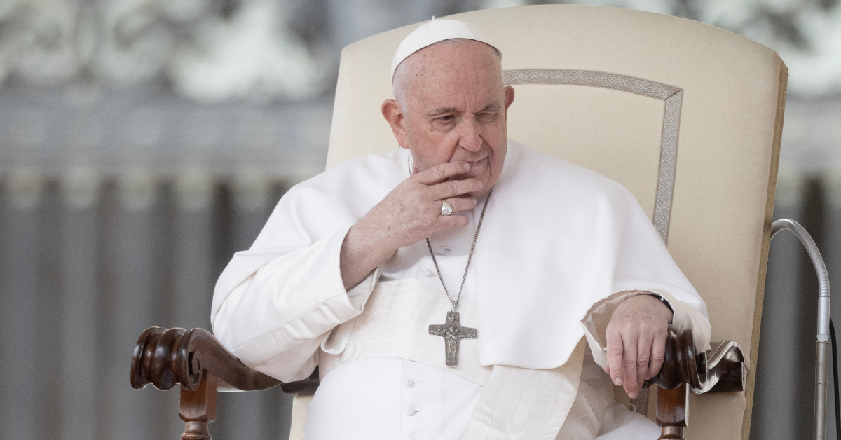 Pope Francis sits in a large cream-colored chair, he has his hand in front of his face and has a contemplative expression.