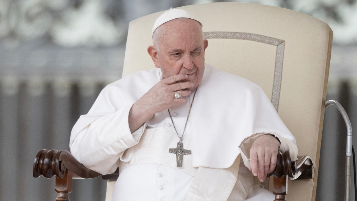 Pope Francis sits in a large cream-colored chair, he has his hand in front of his face and has a contemplative expression.
