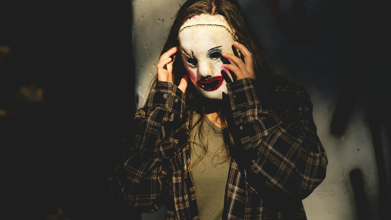 Shot of a long-haired person with a scary mask on. The room behind them is lit is shadows.