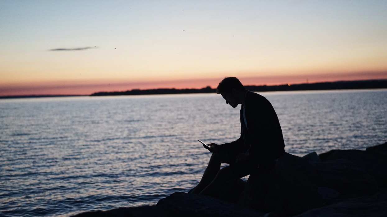 Silhouette of a man sitting by the ocean looking at his phone
